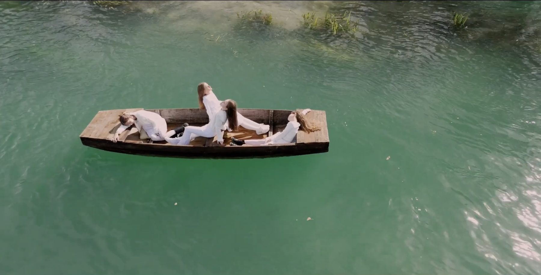 Three young women in white lie in a wooden boat on a green river, still from Somebody Missing
