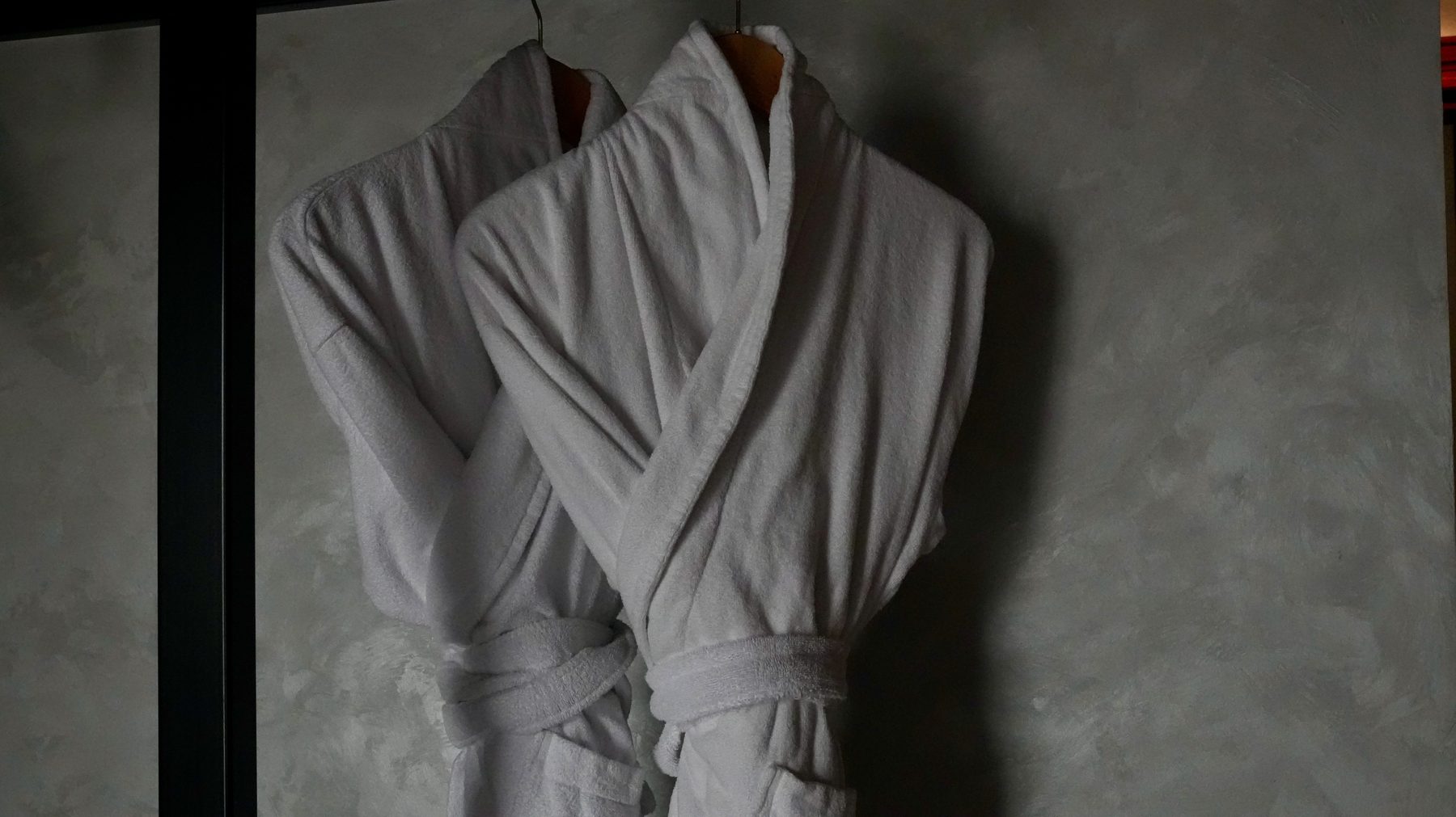 Two white bathrobes on hangers against a plaster wall