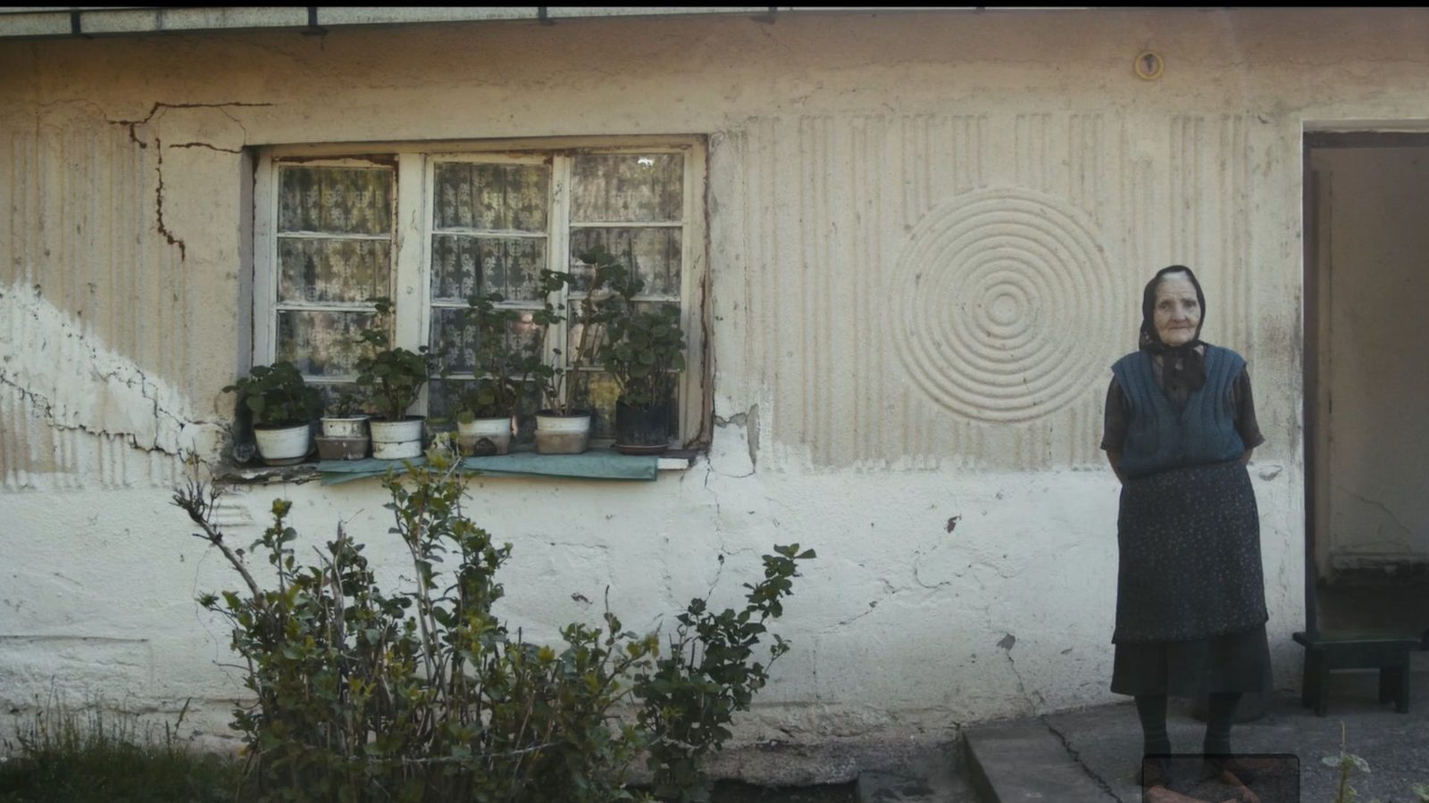 An elderly woman stands by the wall of her old house, still from Ruins, The Last Villagers