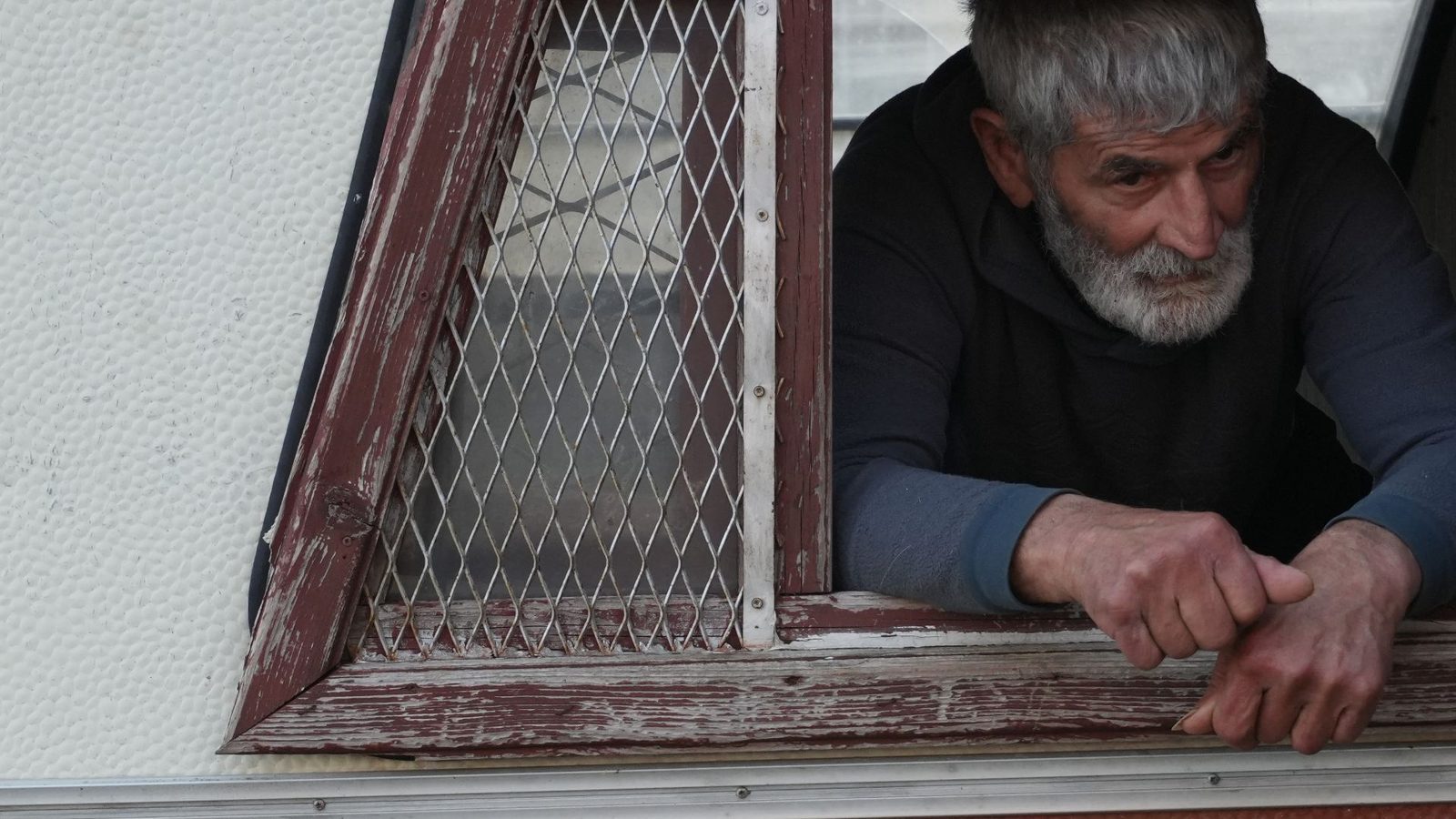 A grey-bearded man leans out of a window with a rusted grille, still from Ruins, The Last Villagers