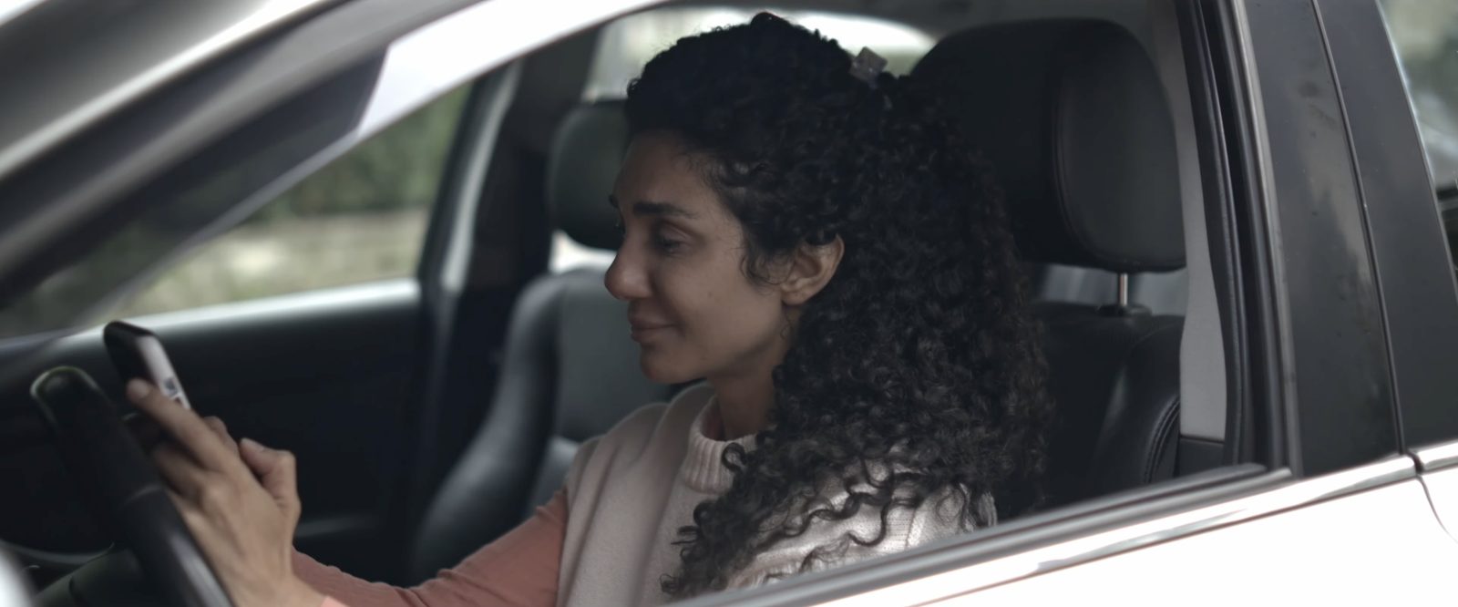 A woman with curly hair smiles at her phone in a car, still from True Love Is Never Forgotten